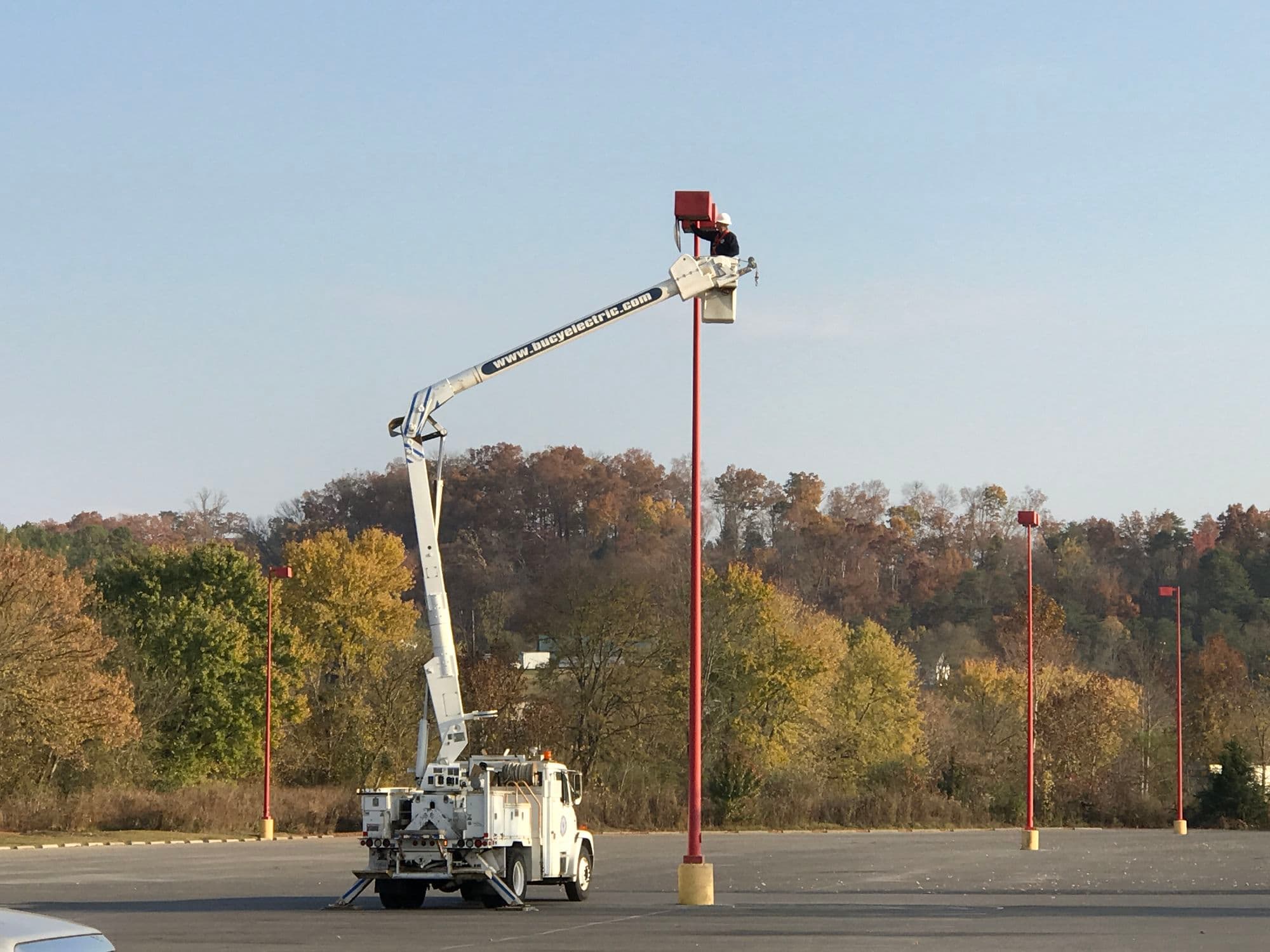 Bucket truck raised to a parking lot light pole with autumn trees in the background